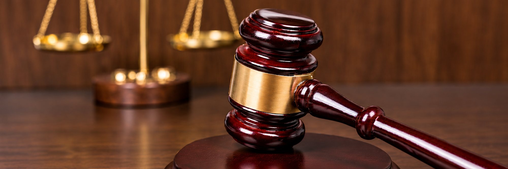 Un marteau de juge en bois sombre à bague dorée posé sur un bureau, avec une balance de la justice floue en arrière-plan.
